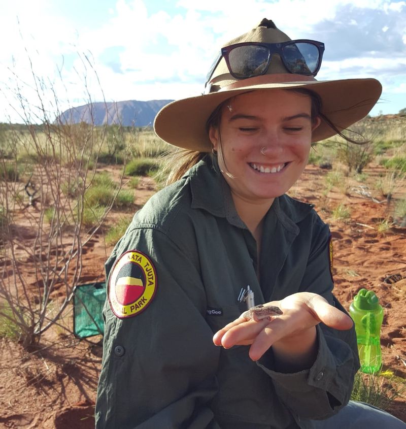 Female ranger holding a small lizard in front of Uluru.