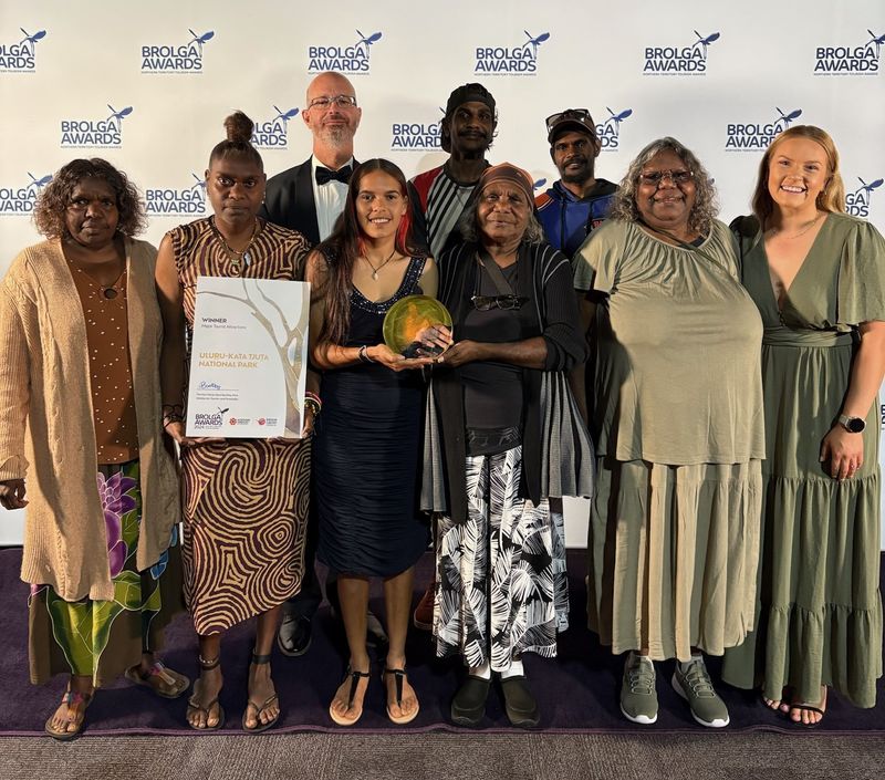 A group of 9 Uluru-Kata Tjuta Anangu Traditional Owners and Parks Australia staff standing in front of a white Brolga Awards sponsorship board, holding a trophy and a certificate.