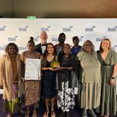 A group of 9 Uluru-Kata Tjuta Anangu Traditional Owners and Parks Australia staff standing in front of a white Brolga Awards sponsorship board, holding a trophy and a certificate