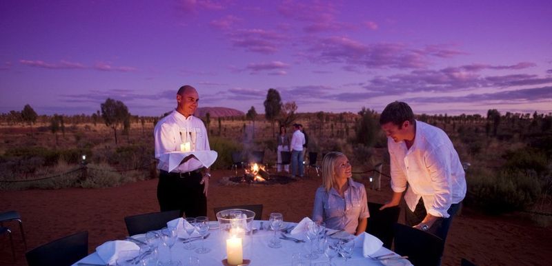 Woman seated at an outdoor table during dusk with Uluru in the backgrounds She is talking to a man as a waiter holds two glasses of sparkling wine..