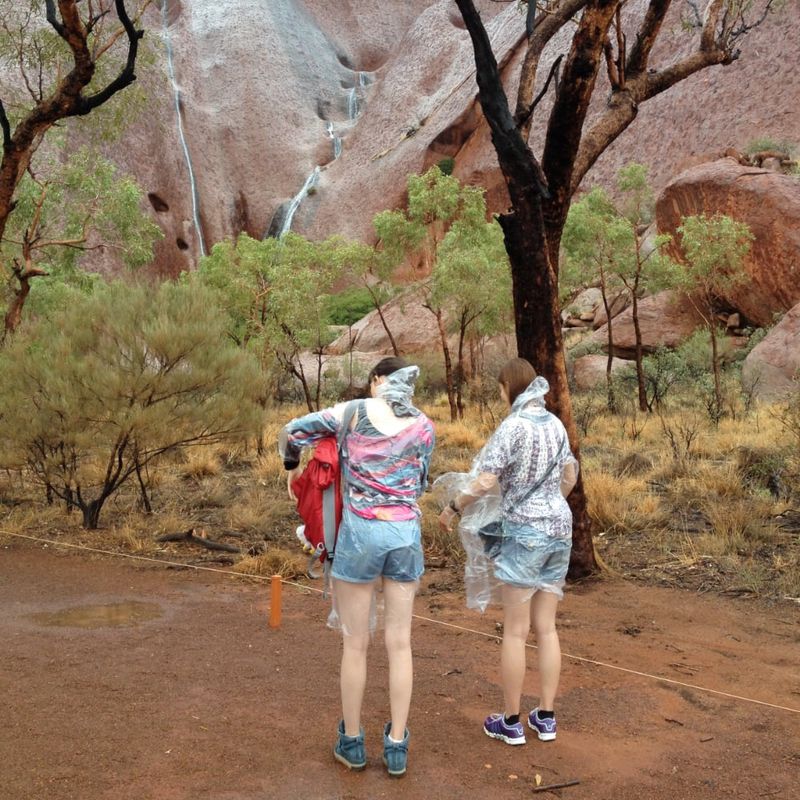 Two women wearing rain ponchos in front of an Uluru rock face.